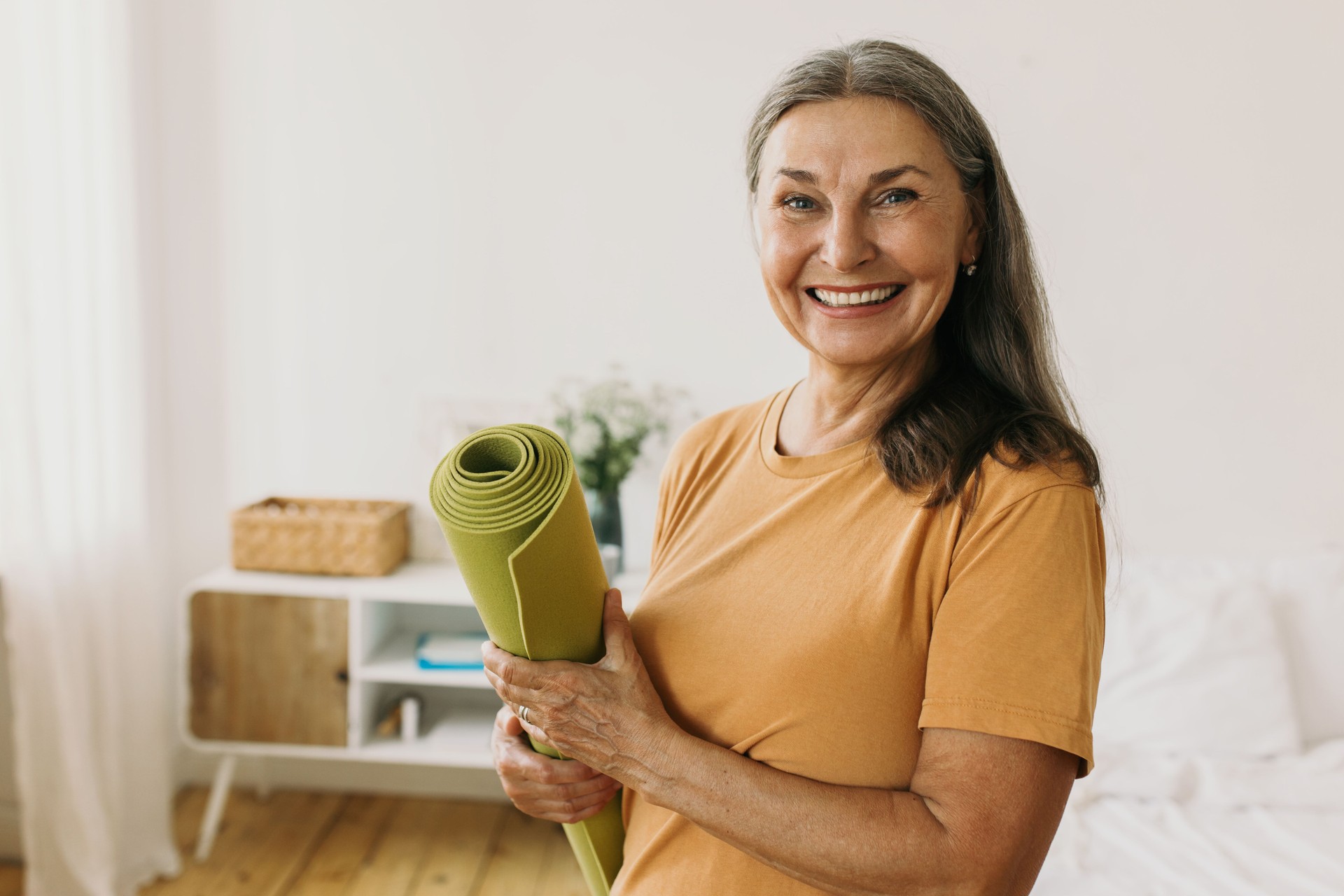 Happy cheerful joyful mature 60-years-old sporty woman standing against bed at her apartment with rolled green mat in hands, waiting with anticipation beginning of daily physical workout