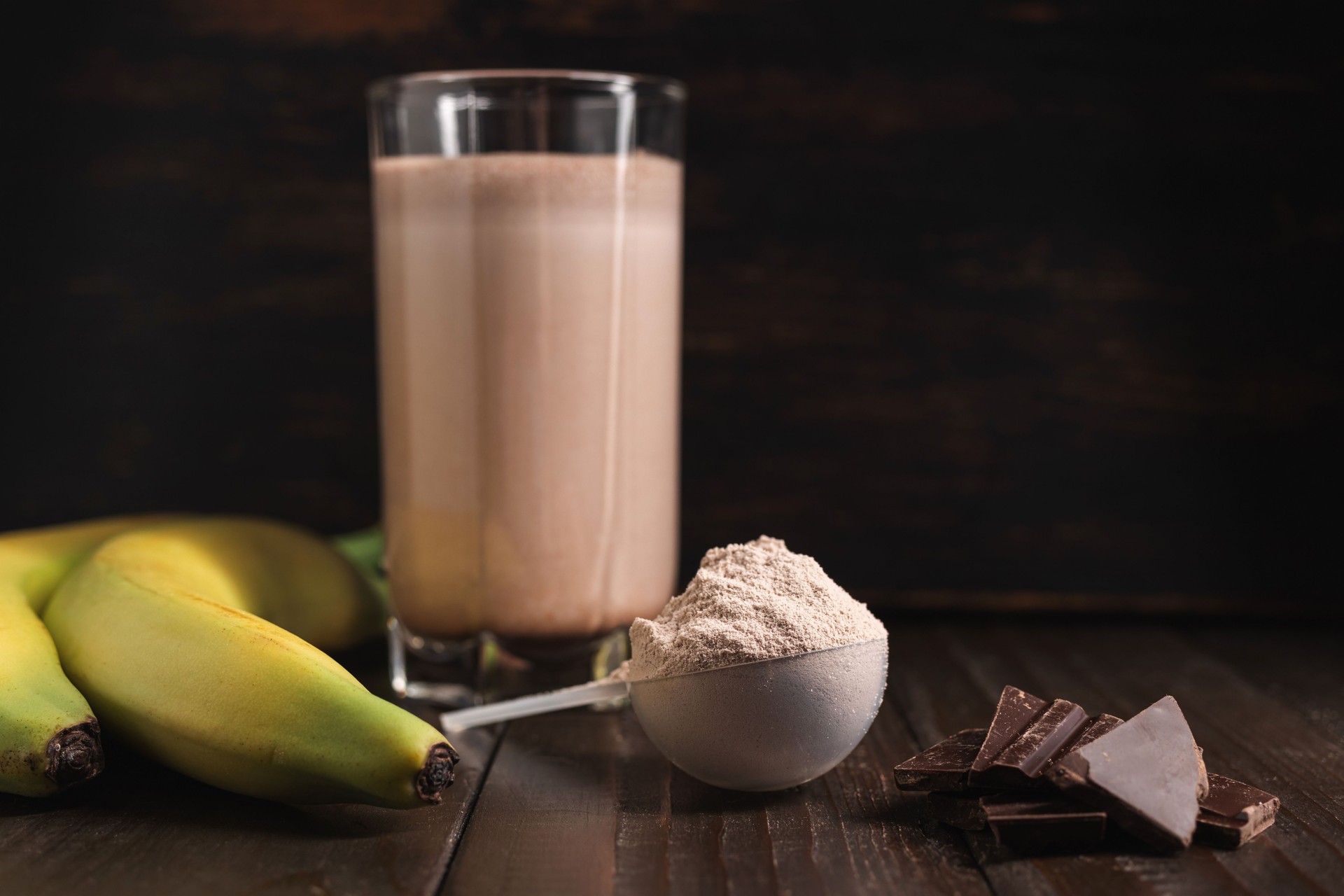 Plastic measuring spoon with whey protein powder, chocolate milkshake cocktail in a glass, blended protein drink and banana fruit on a dark wooden background